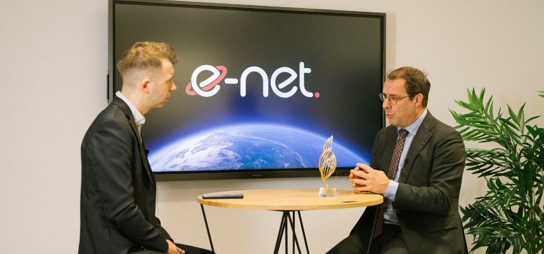 Interview dans les locaux e-net, Sacha Peiffer et David Clarinval en discussion, assis autour d’une petite table, devant un écran affichant le logo de l’entreprise et le trophée de l’innovation.