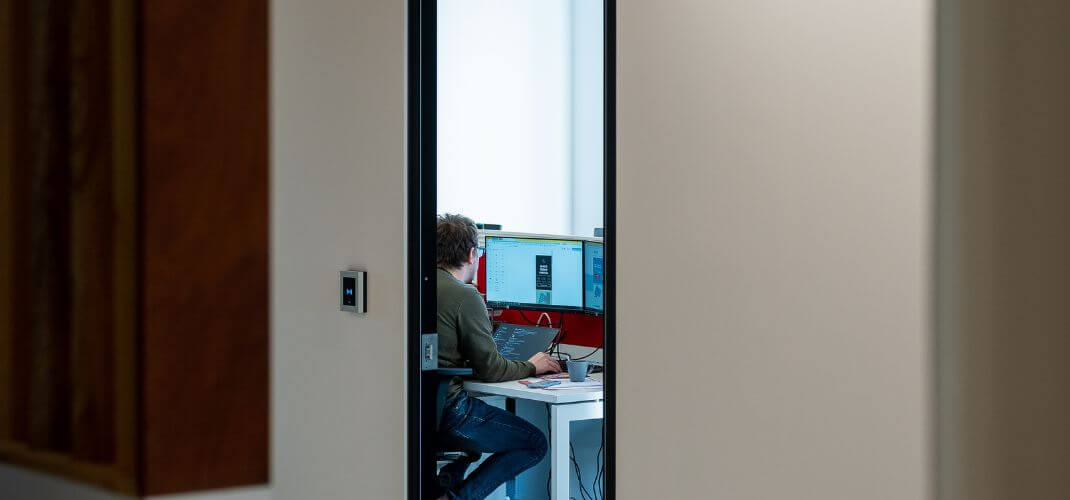 Vue à travers l’embrasure d’une porte dans les locaux e-net, montrant une personne travaillant à un bureau devant plusieurs écrans.